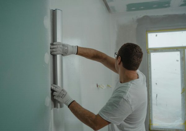 A man in a white shirt and gloves using a metal ruler for indoor wall renovation work.